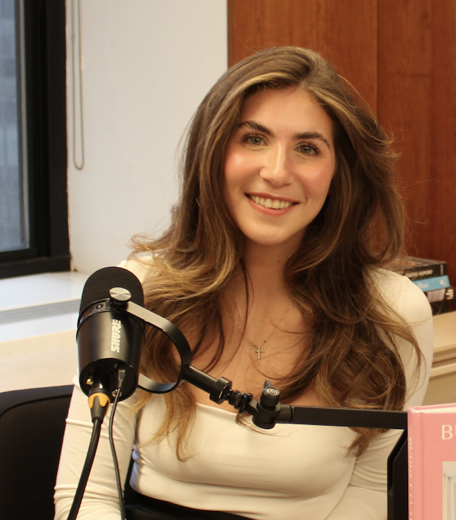 A woman with long brown hair, wearing a white top, sits at a desk with a microphone in front of her, smiling at the camera in a podcast or interview setting.